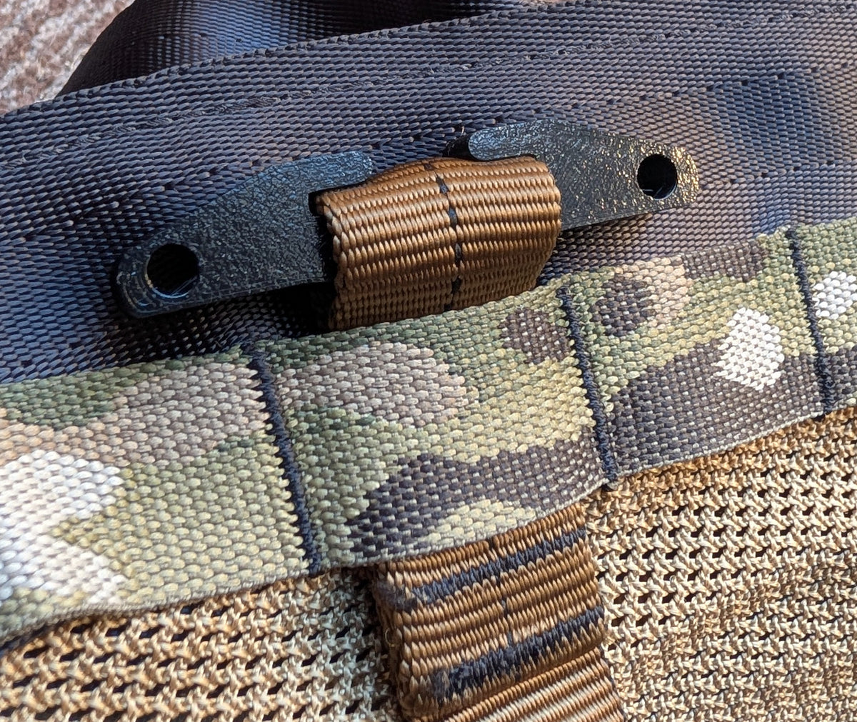 Close-up detail of a tactical bag showing a camouflage webbing strap with a tan plastic buckle, attached to a dark gray webbed loop over a tan mesh fabric.