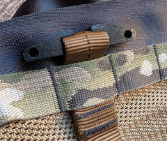 Close-up detail of a tactical bag showing a camouflage webbing strap with a tan plastic buckle, attached to a dark gray webbed loop over a tan mesh fabric.